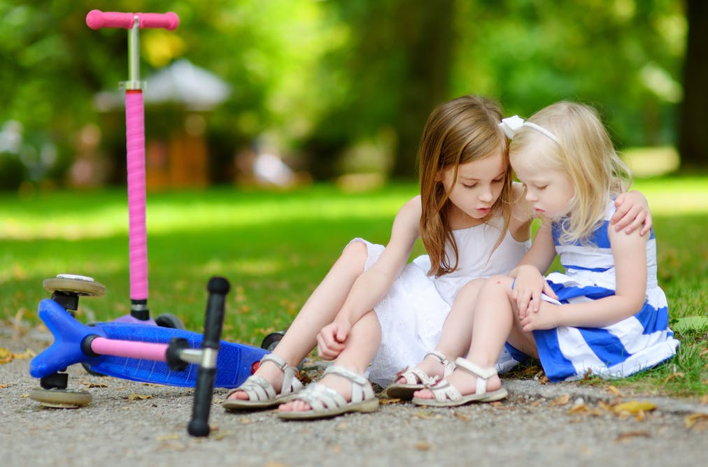 Two young girls sitting on the grass in a park with their scooters nearby.