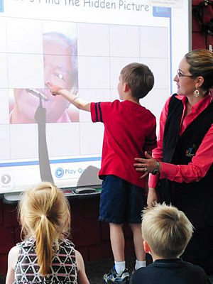 Child solving puzzle on interactive whiteboard with teacher and classmates watching