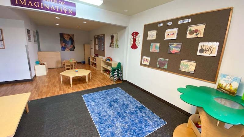 Colorful preschool classroom with play area tables and an art display board on the wall.