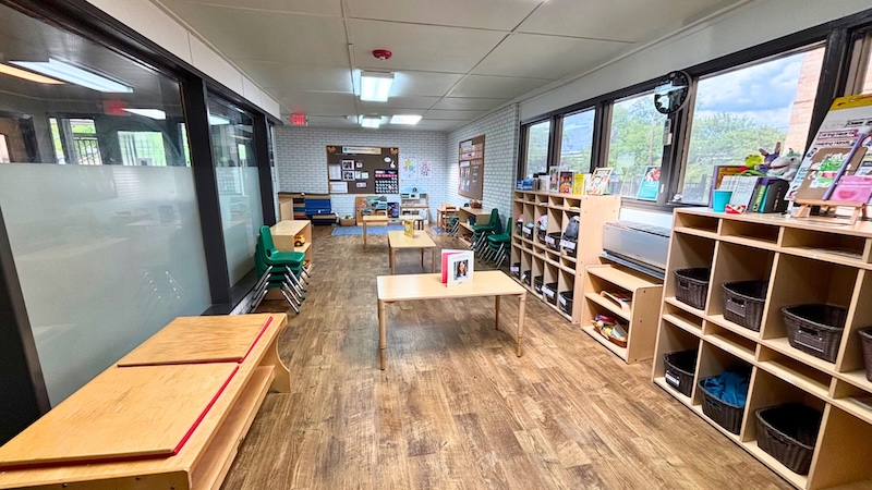 Spacious preschool classroom with wooden floors, tables, shelves, and large windows.