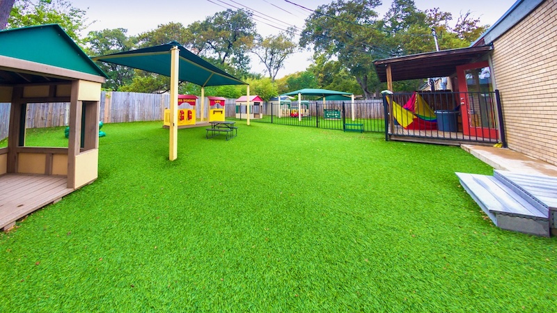 Outdoor preschool playground with playhouses and shade structures on artificial grass.