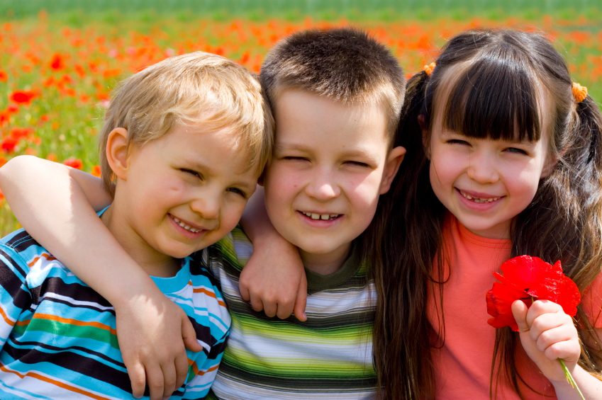 Three smiling children embracing in a field of red flowers on a sunny day