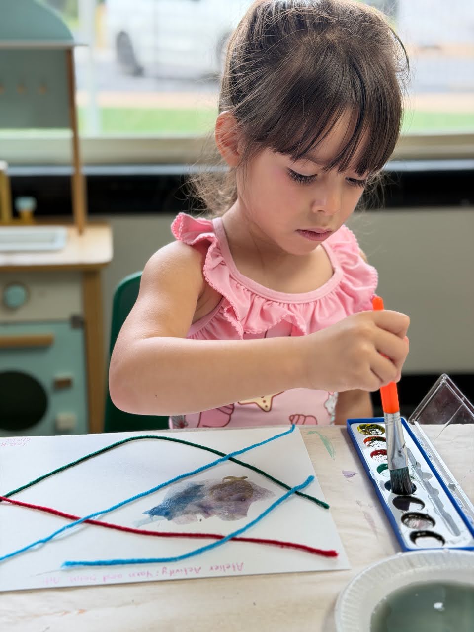 Young girl painting with watercolors in a classroom setting wearing a pink dress focused on her artwork