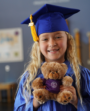 Young graduate in blue cap and gown holding a teddy bear, smiling proudly indoors