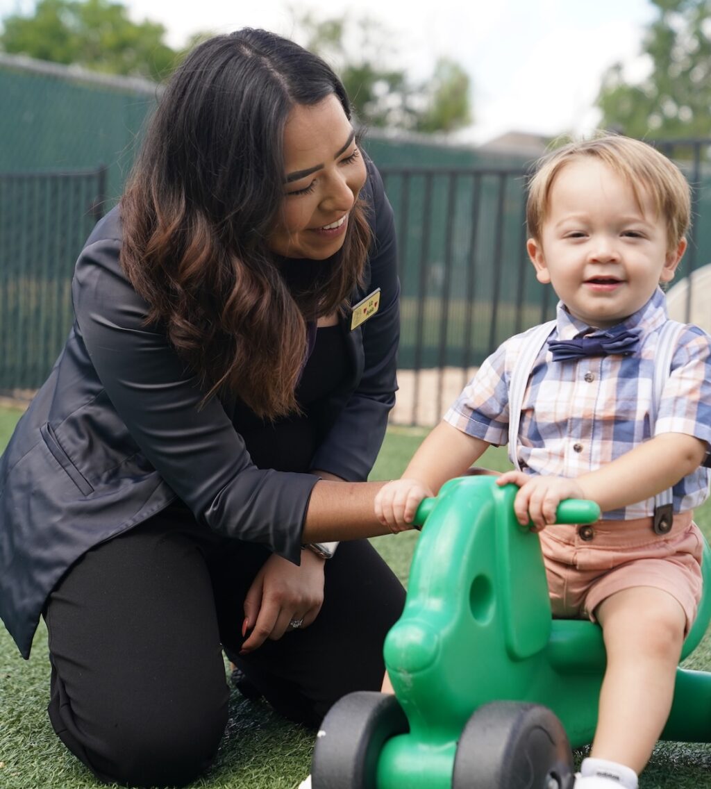 Teacher smiling with a young child on a green toy in a playground setting
