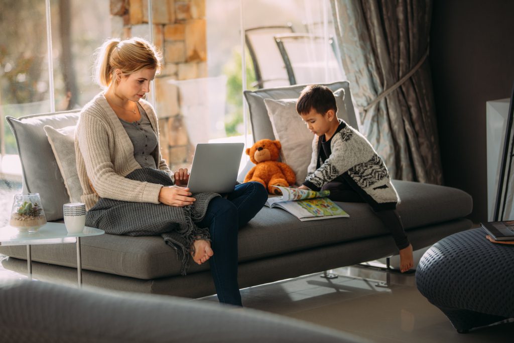 Woman working on laptop while child reads a book on sofa in cozy living room