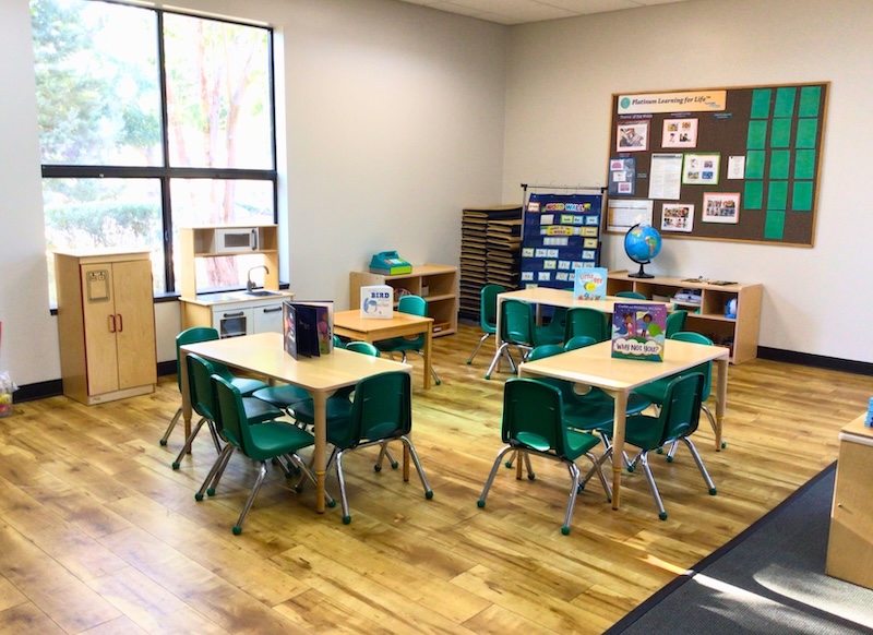 Colorful preschool classroom with small tables and chairs, books, and educational displays set up for young learners.