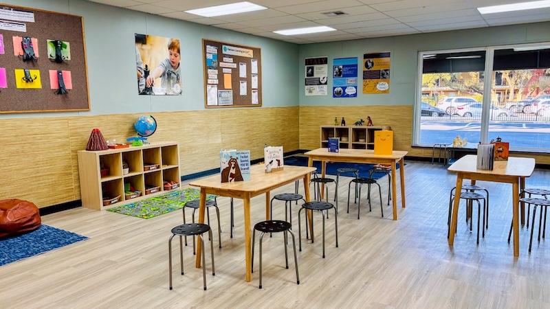 Bright, organized classroom with tables, chairs, educational displays, and books on natural wooden shelves.