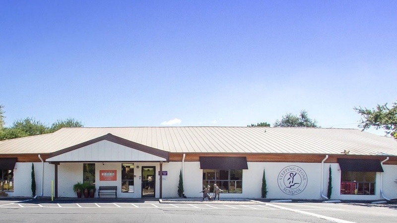 Single-story school building with awnings on a sunny day, featuring a large logo on the wall.
