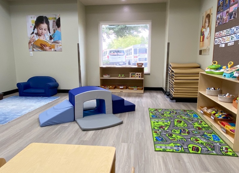 Bright daycare room with play area, books, and toys, featuring blue chairs and educational wall displays.