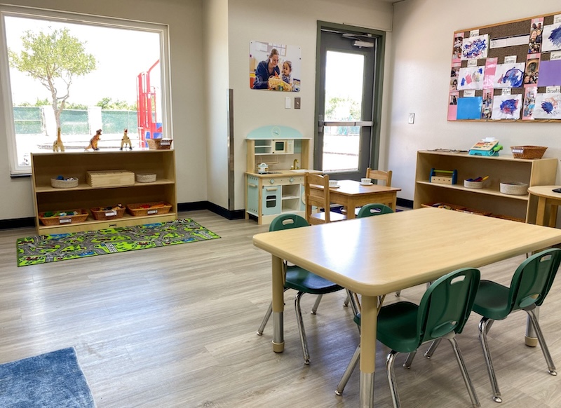 Bright, organized daycare classroom with tables, chairs, shelves, and a play kitchen by a large window.