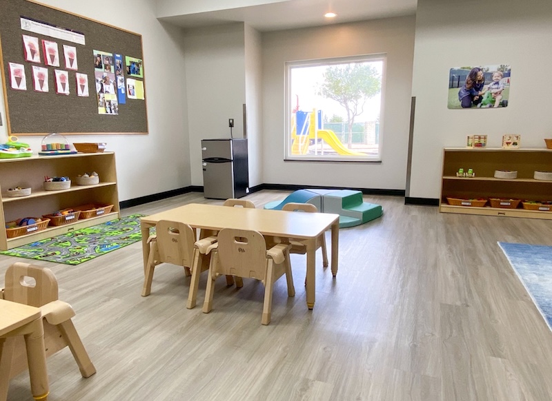 Bright preschool classroom with small tables chairs shelves and a window view of a playground slide