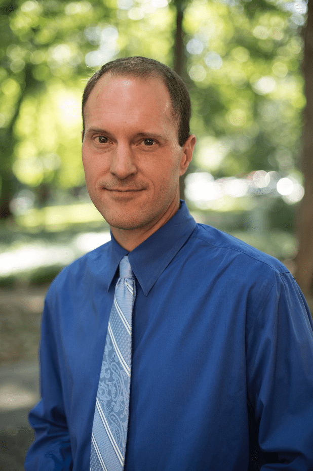 Man in blue shirt and tie standing outdoors with blurred green foliage background