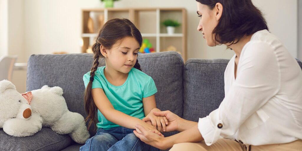 Woman comforting a young girl on a sofa in a cozy living room with a teddy bear nearby.
