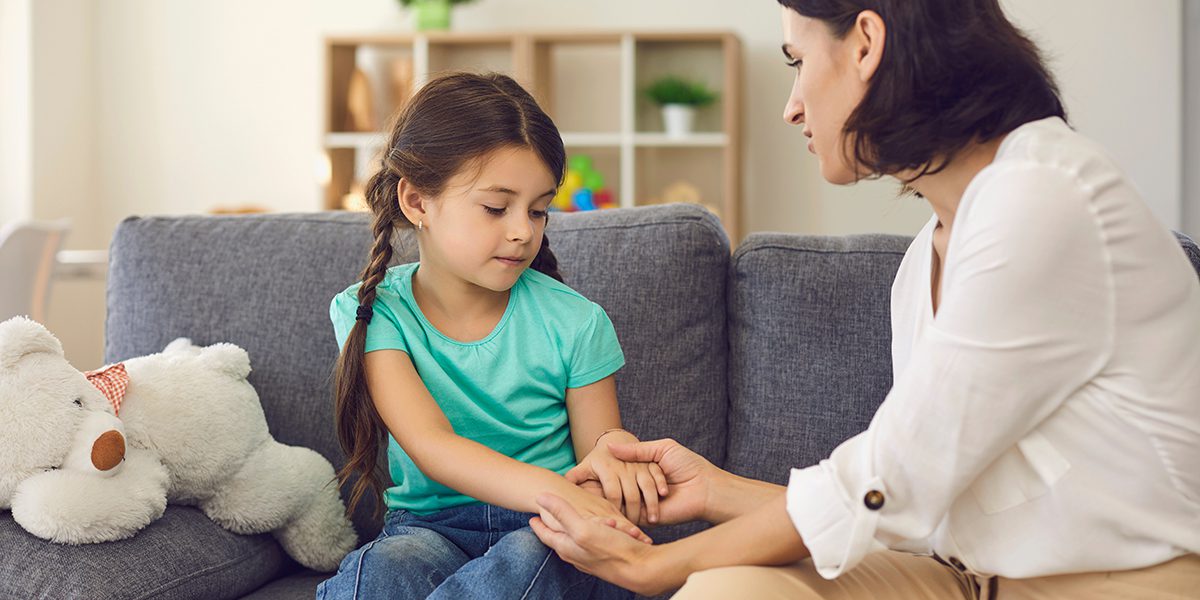 Woman comforting a young girl on a sofa in a cozy living room with a teddy bear nearby.