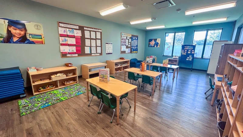 Bright classroom with tables, chairs, educational posters, and wooden shelves for young children