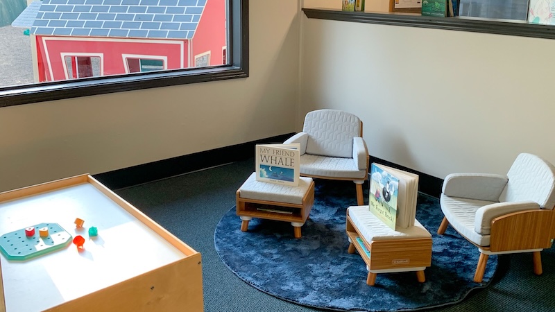 Cozy kids reading nook with chairs and books near a sunlit window displaying a quaint red house outside