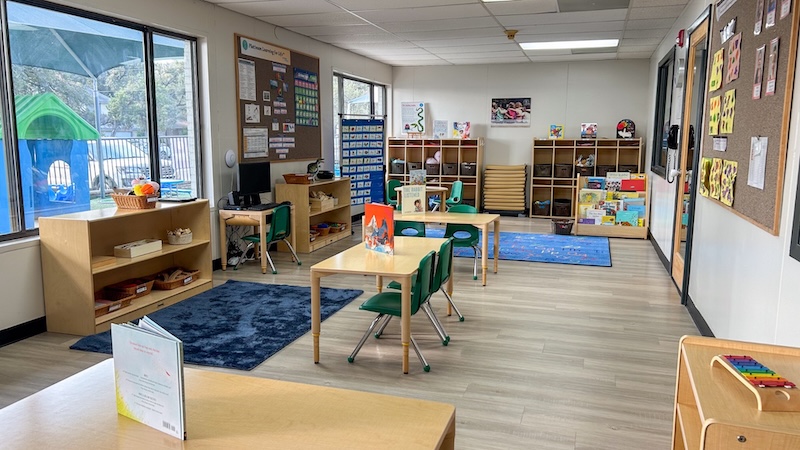 Bright preschool classroom with tables; shelves filled with books and toys; large windows and colorful decor.