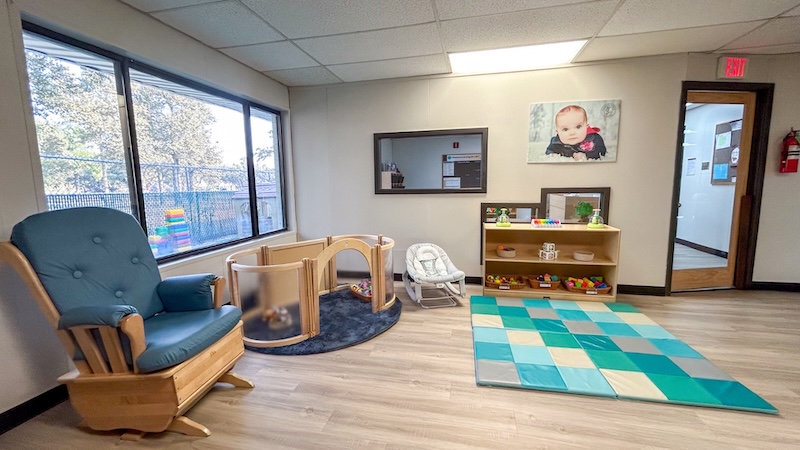 Bright daycare room with rocking chair, toys, and play mat against large windows in a well-lit space.