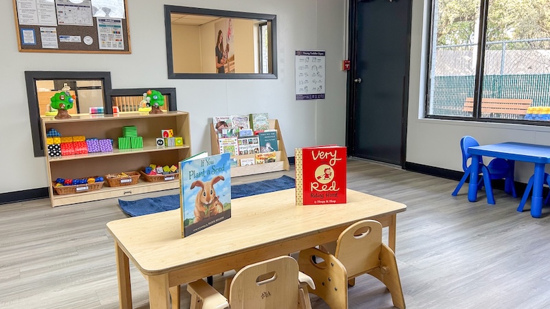Colorful preschool classroom with a wooden table, books, toys, and educational materials arranged neatly.
