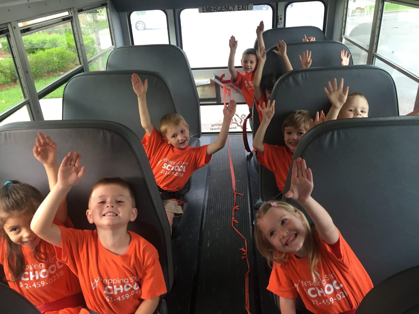 Kids in orange shirts smiling and raising hands on a school bus.
