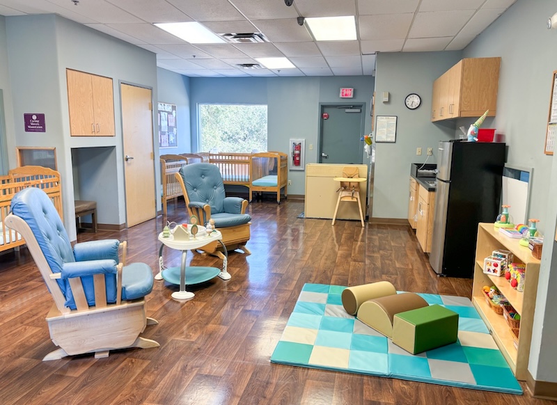 Daycare room with cribs, rocking chairs, play mat, and toys on wooden floor.