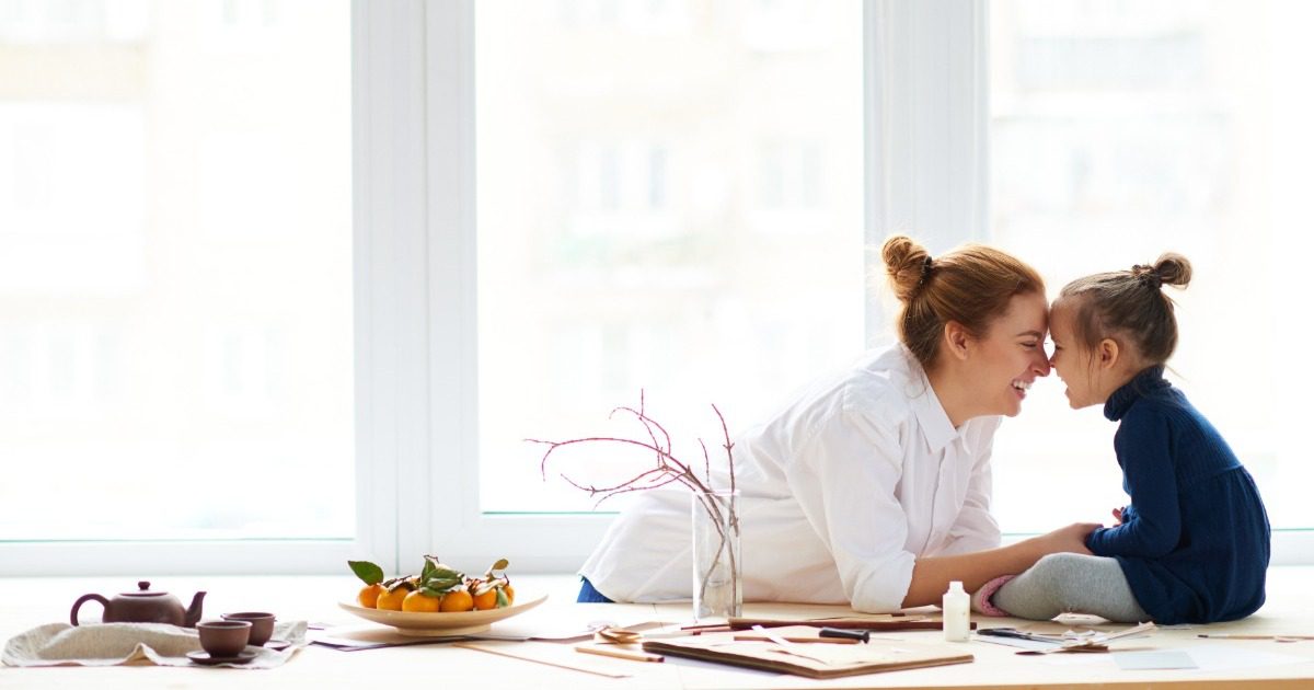 Mother and daughter play together at a bright table near a large window.