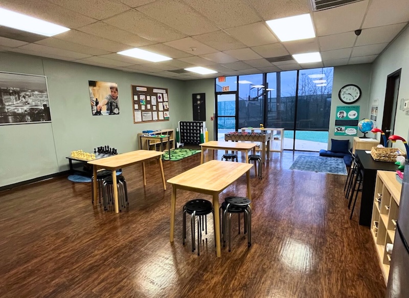 Bright classroom with tables, stools, chess sets, and educational displays on wooden flooring with large windows