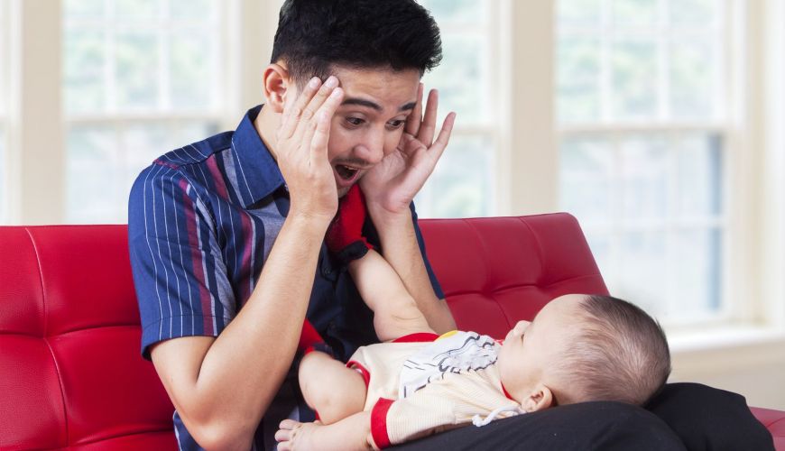Man making funny faces at a baby on a red couch in a bright room