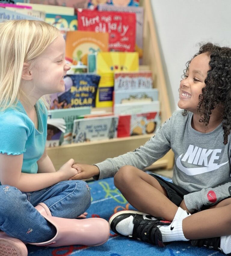 Two children sitting and smiling while holding hands in a colorful library setting