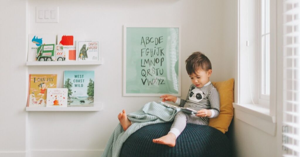 Toddler reading a book in a cozy, well-lit nook with bookshelves and alphabet poster.