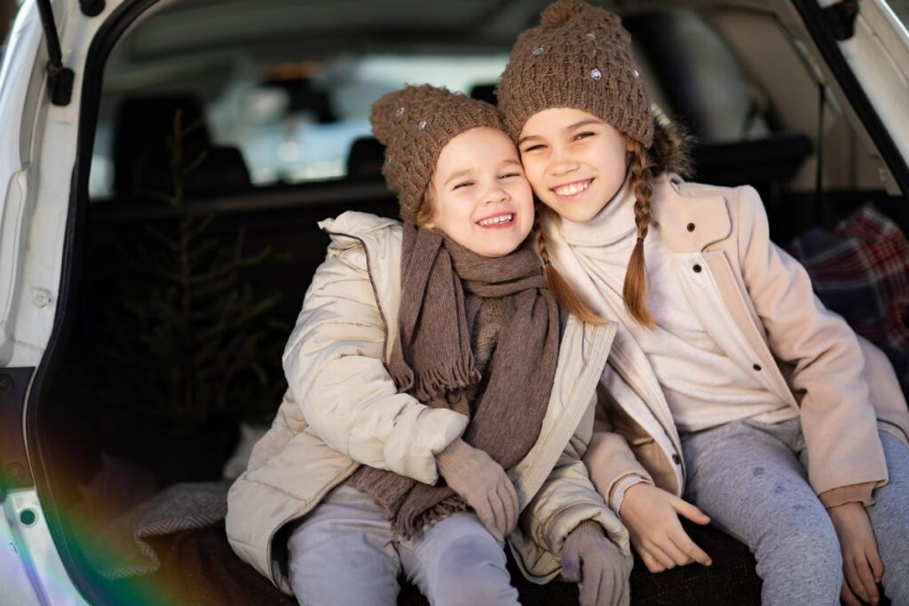 Two smiling children in winter coats and hats sit in the back of a car enjoying a cold day.