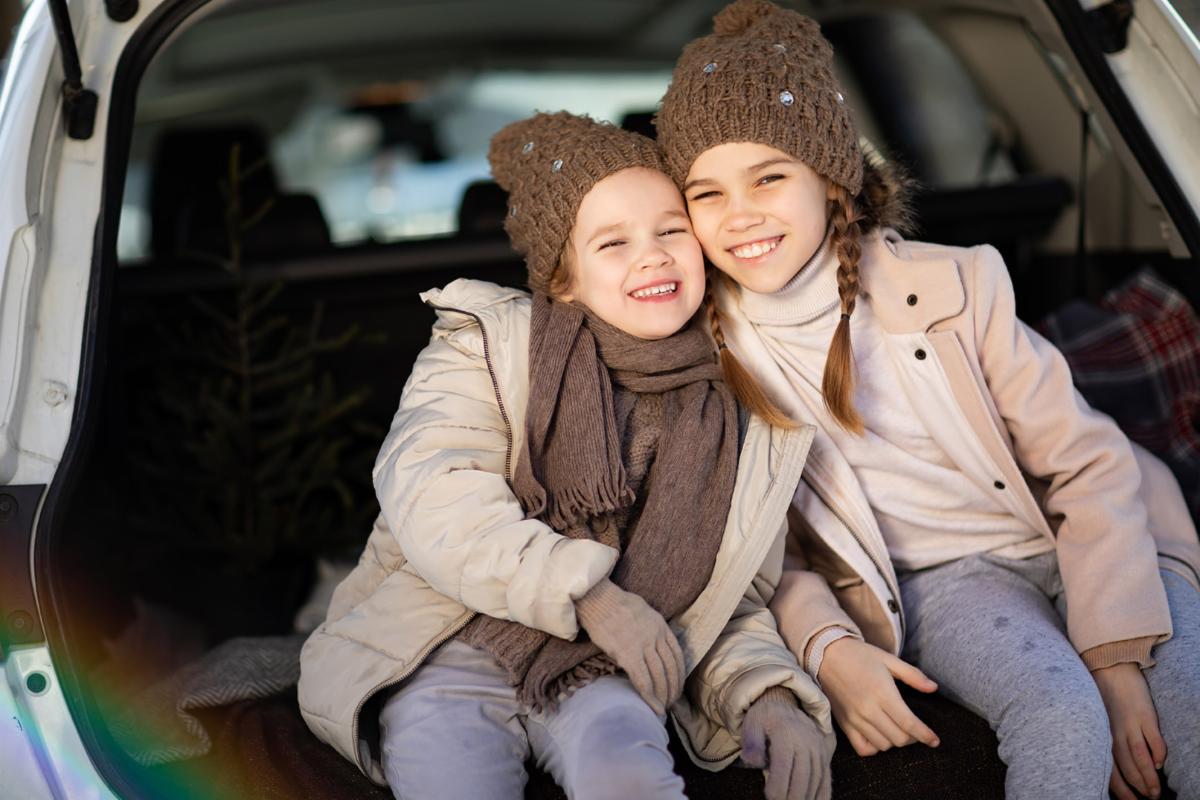 Two smiling children in winter coats and hats sit in the back of a car enjoying a cold day.