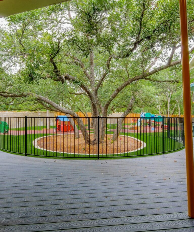 Playground with large tree surrounded by fence and play equipment on artificial turf.