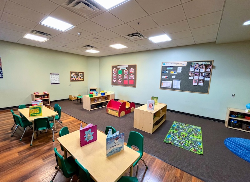Colorful preschool classroom with tables, chairs, books, and play area on a wooden floor with bright lighting.