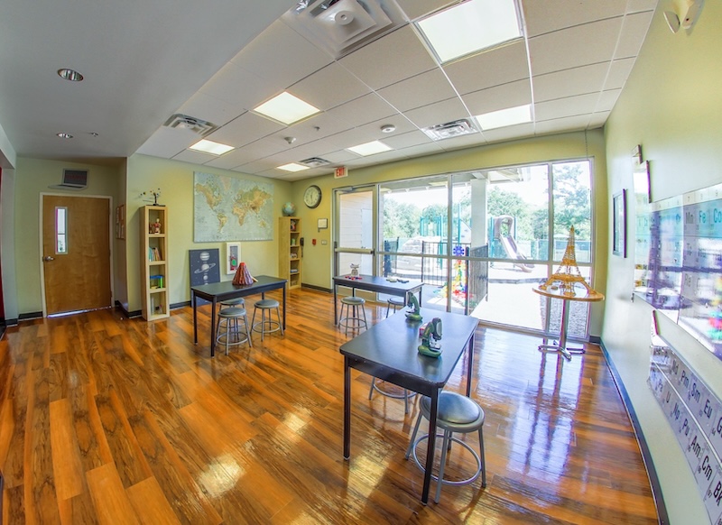 Bright classroom with wooden floors and large windows overlooking a playground, featuring desks and educational decor.