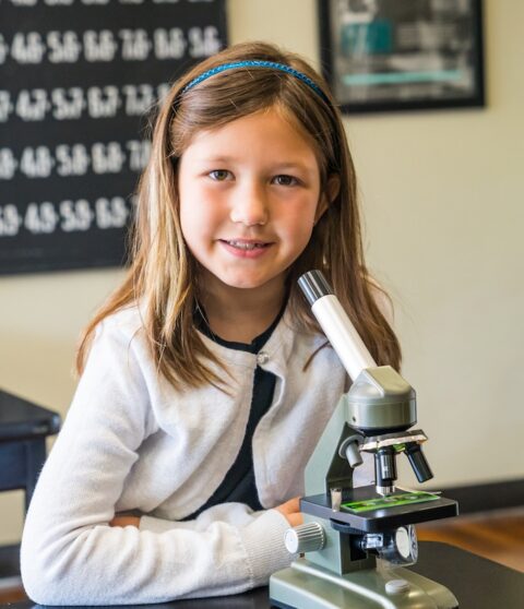 Young girl smiling at desk with a microscope and classroom background.