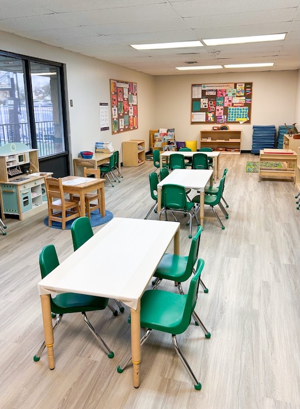 Bright preschool classroom with small tables and green chairs, educational posters, and play areas.