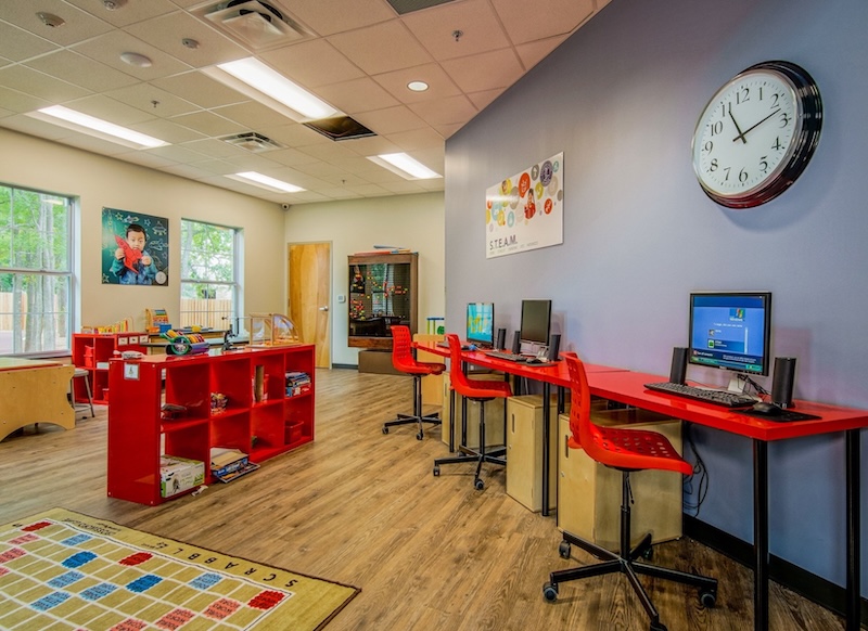 Colorful preschool classroom with computers, red furniture, educational toys, and a wall clock