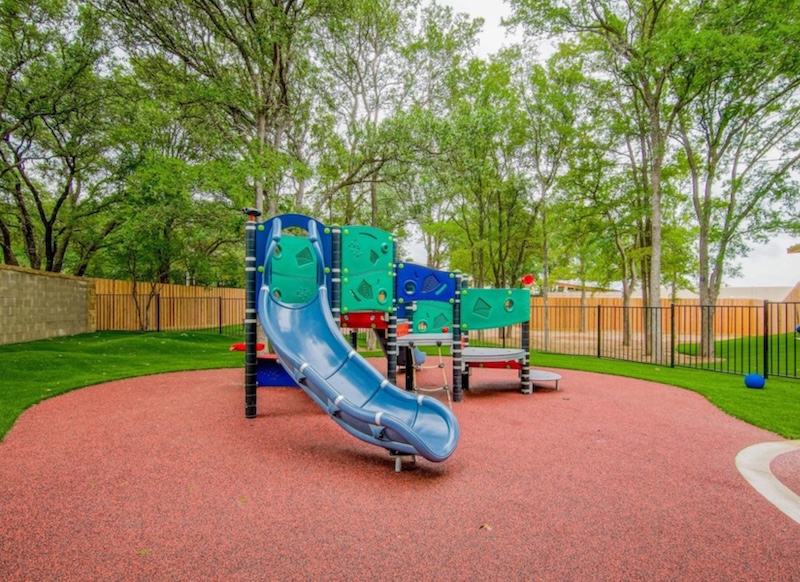 Playground with slide and climbing structures on a soft surface surrounded by trees and a fence