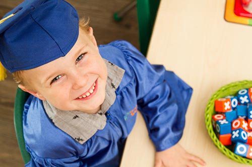 Child in blue graduation cap and gown smiling next to colorful blocks on a table