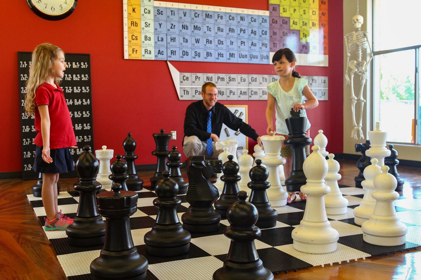 Children playing with large chess pieces in classroom with periodic table on wall