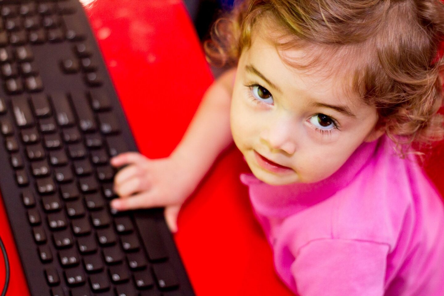 Toddler in pink shirt using a keyboard on a red table.