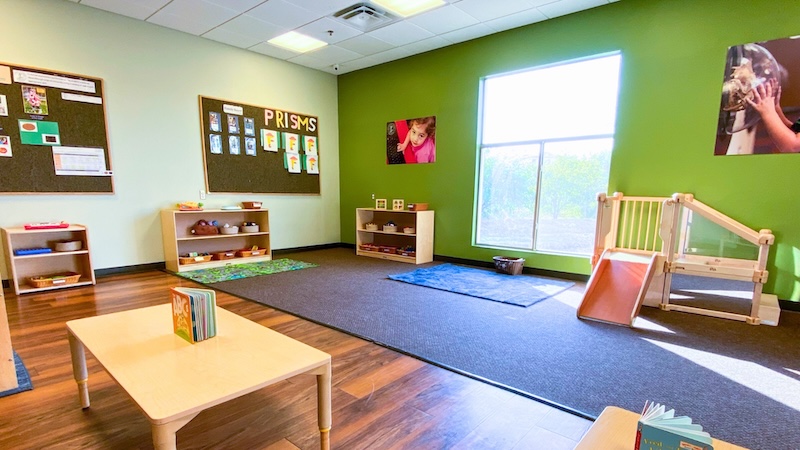 Bright preschool classroom with learning materials, small slide, and large window on green walls.