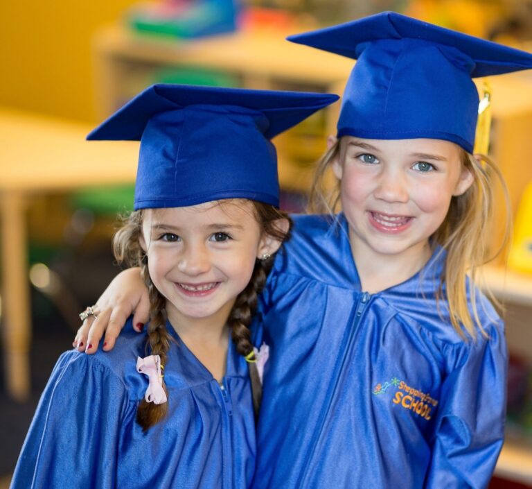 Two young girls in blue graduation gowns smiling at a preschool ceremony