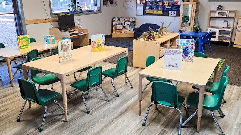 Bright classroom with tables, green chairs, and children's books on display.