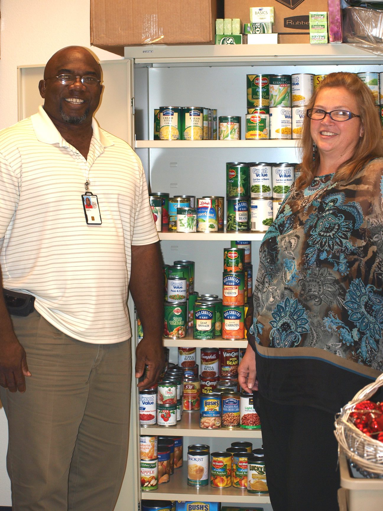 Two people smiling near a well-stocked pantry with various canned goods.